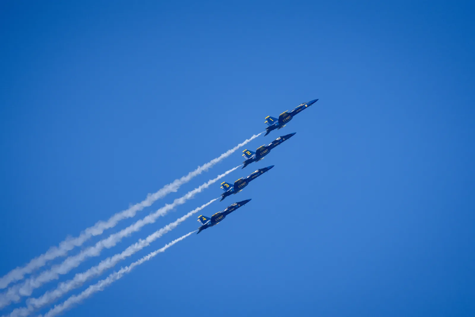 Blue Angels jets fly in a diagonal line with smoke trails across the sky.