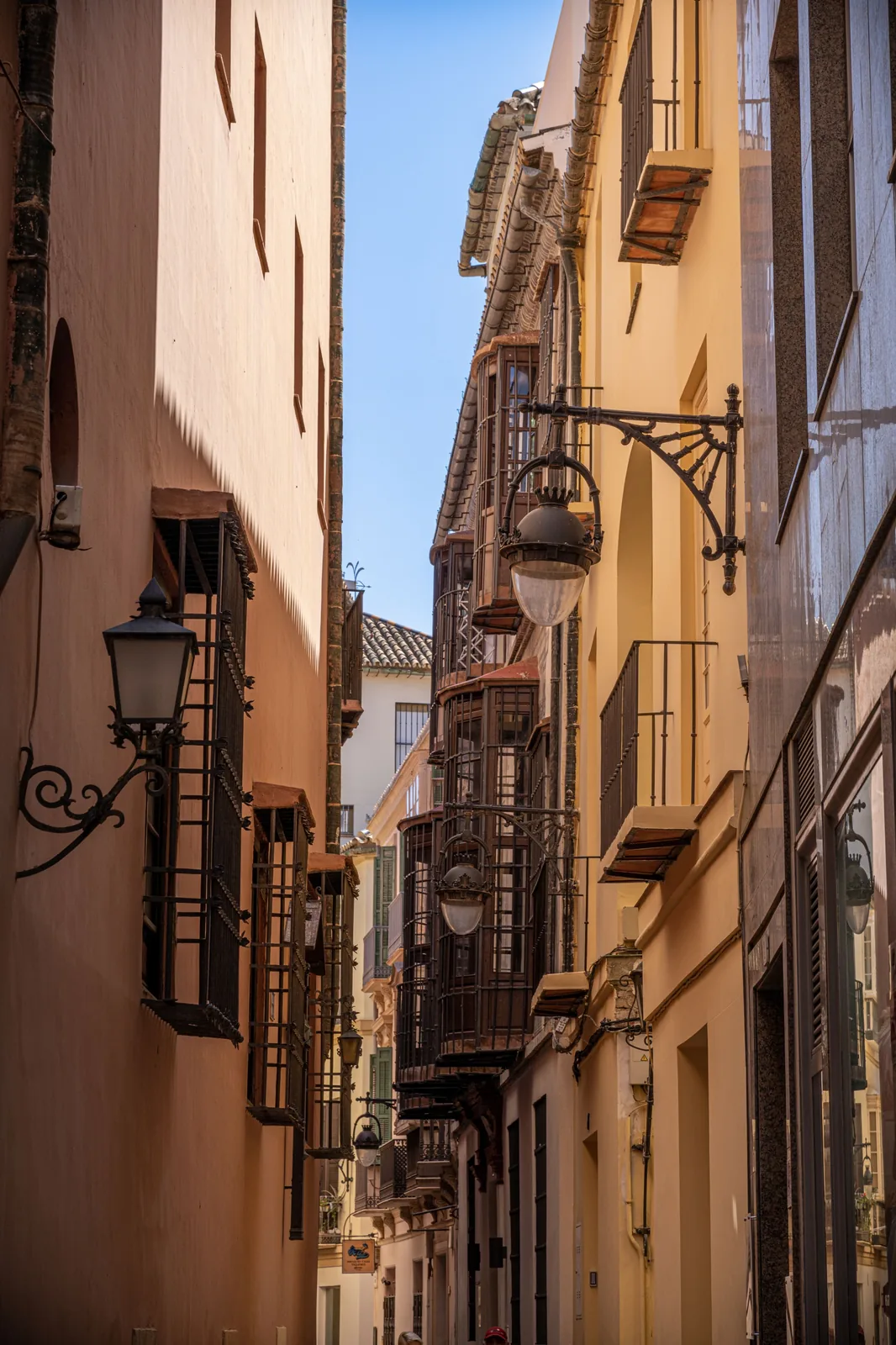 Narrow street lined with colorful buildings, enclosed balconies, barred windows, and ornate hanging lamps beneath a strip of blue sky.