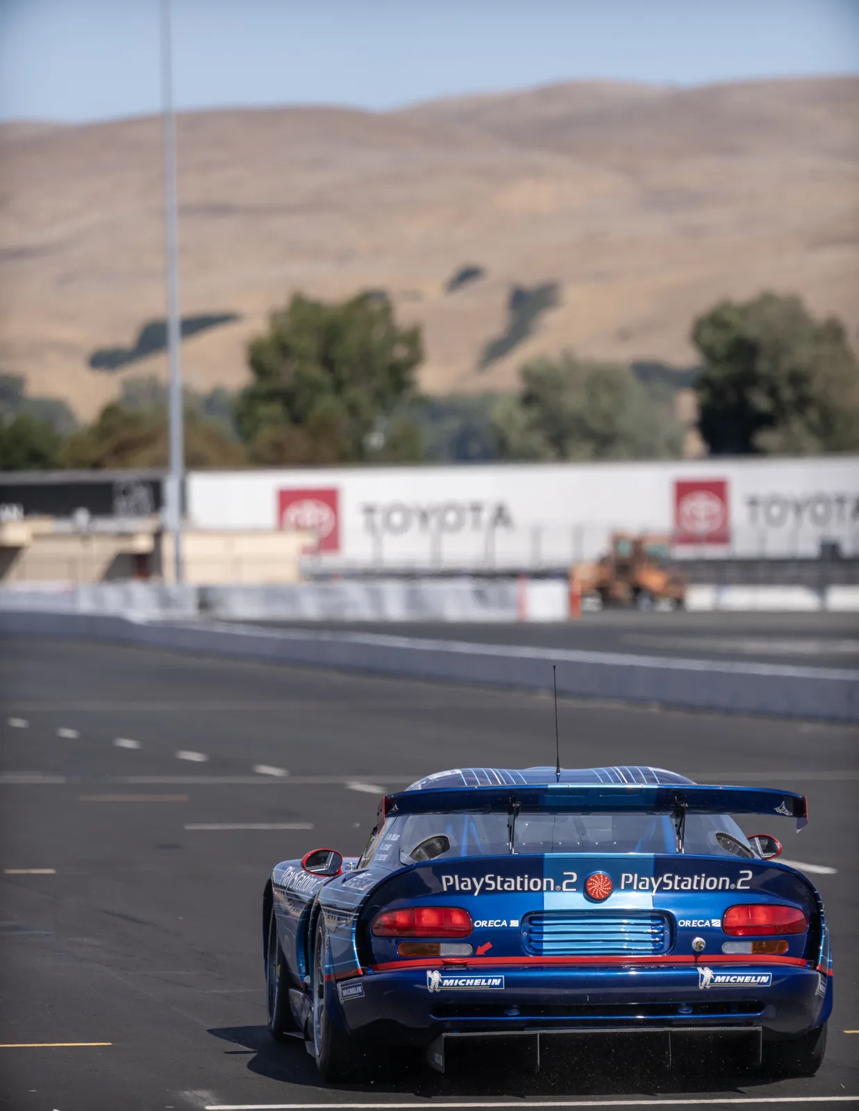 Rear view of a blue race car with PlayStation 2 and Michelin branding on a paved track, with blurred hills and barriers behind it.