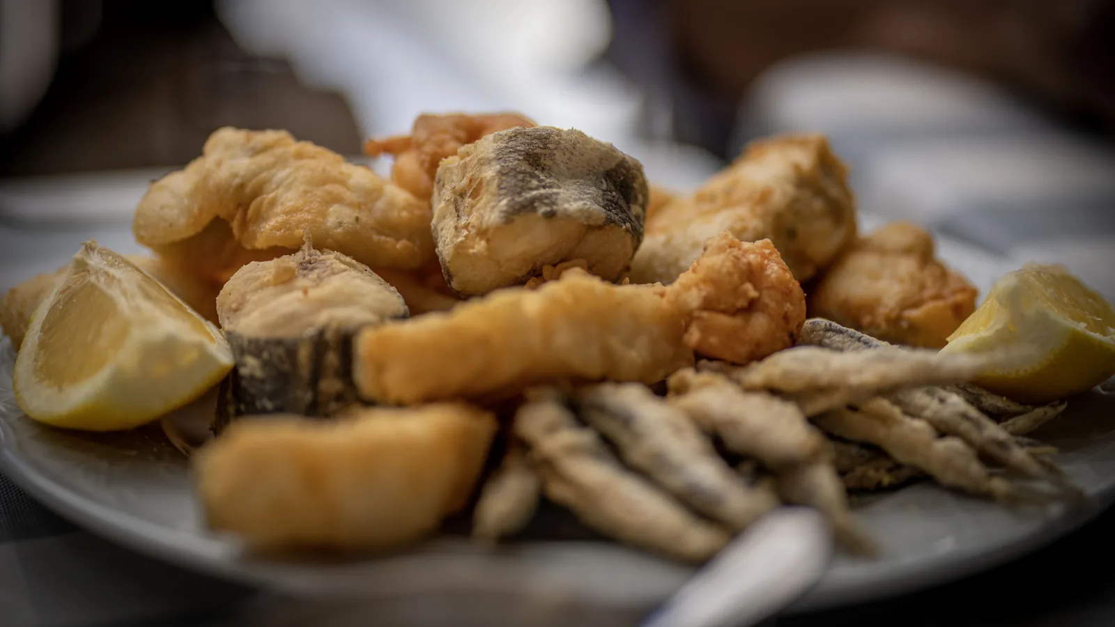 A shallow plate holds assorted battered fried seafood and small whole fish, with lemon wedges at the edges and a blurred dining setting behind.