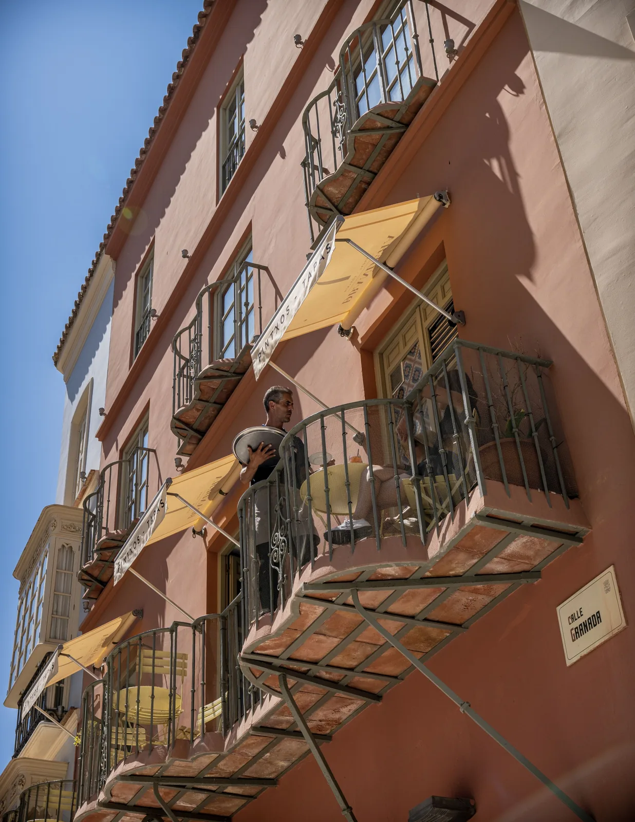 Man carrying a large round tray stands on a balcony of a pink building with iron railings and yellow awnings.