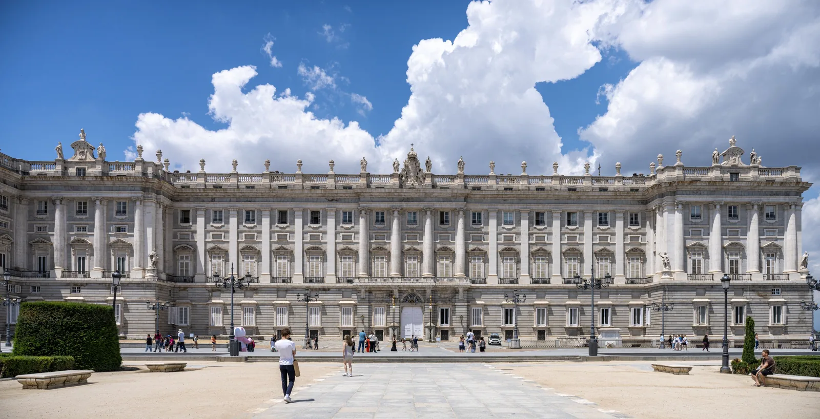 Broad symmetrical palace facade faces an open plaza where visitors walk among clipped hedges and lamps beneath large white clouds.
