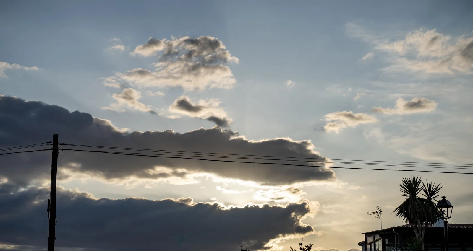 Layered clouds part around bright low sunlight above rooftops, crossed by utility wires and a pole, with an antenna and palm-like plant at right.