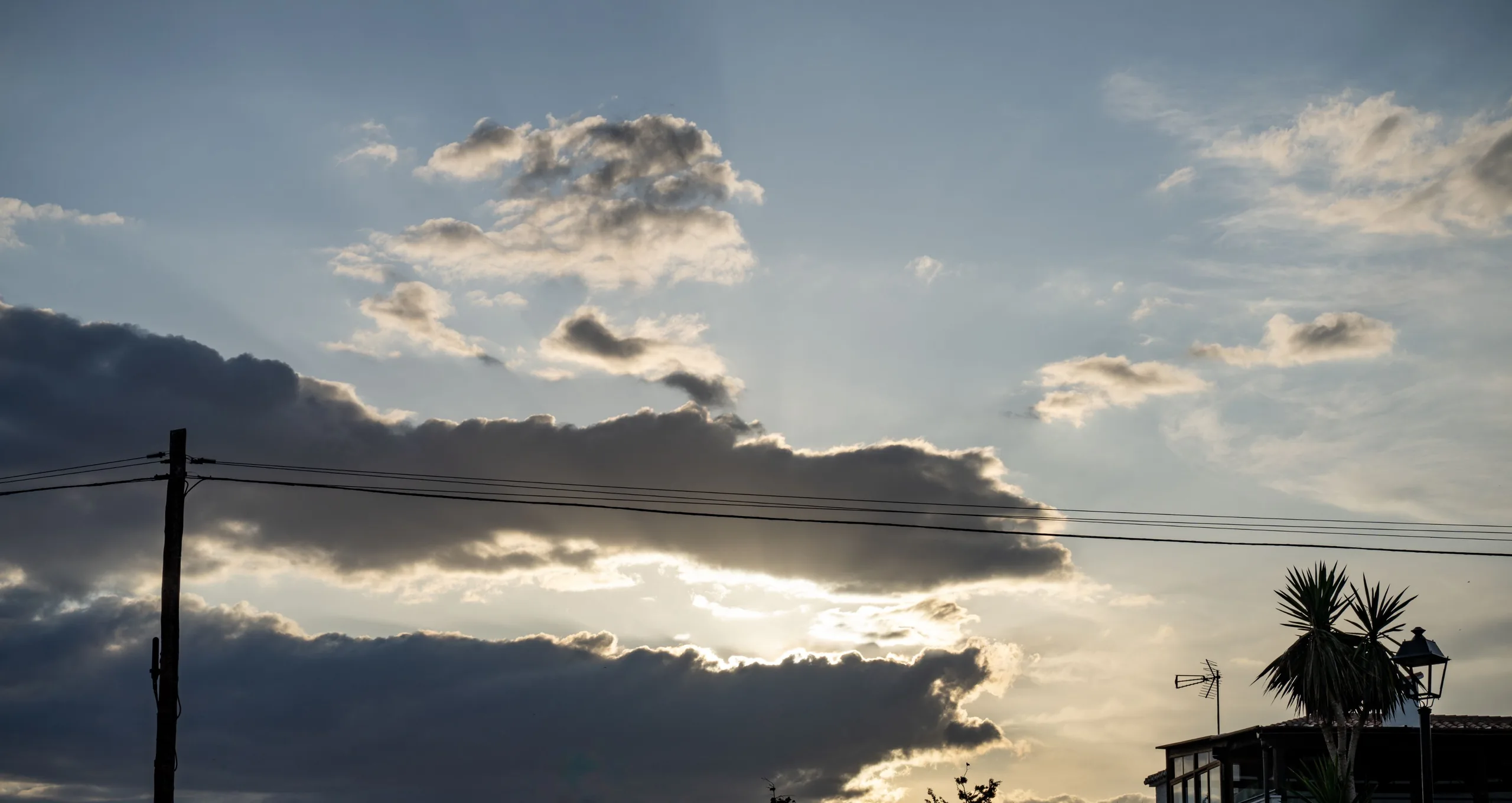 Layered clouds part around bright low sunlight above rooftops, crossed by utility wires and a pole, with an antenna and palm-like plant at right.