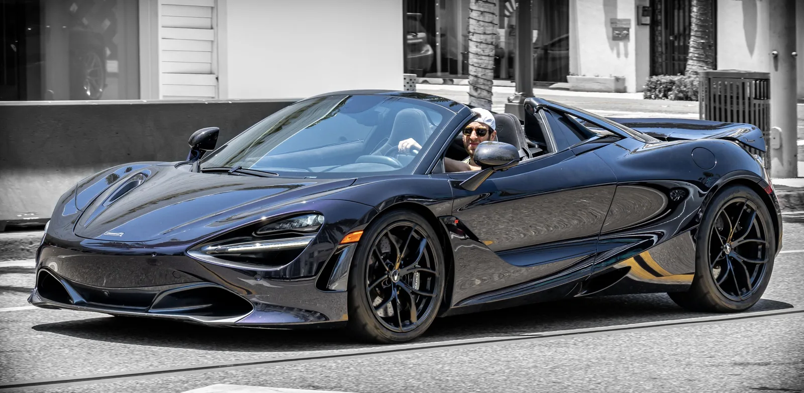 Dark convertible sports car with a driver visible passes along a city street, shown in a side-front view with storefronts behind it.