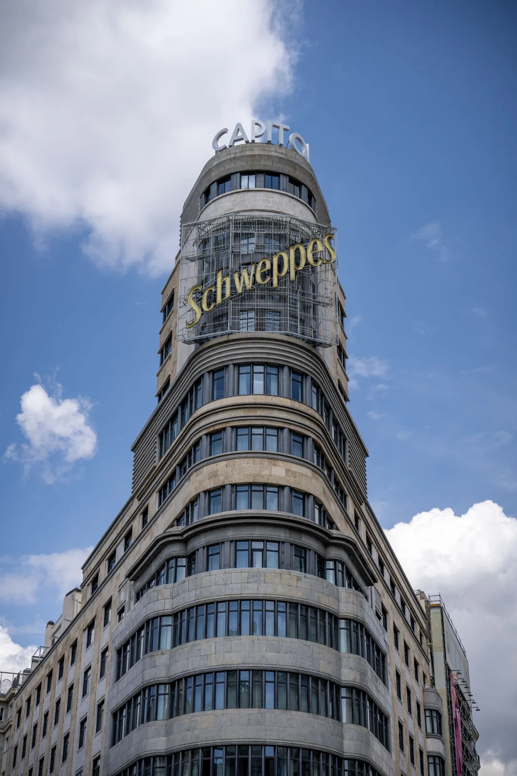 Rounded multi-story stone building with curved window bands and large CAPITOL and Schweppes signs stands against a partly cloudy sky.