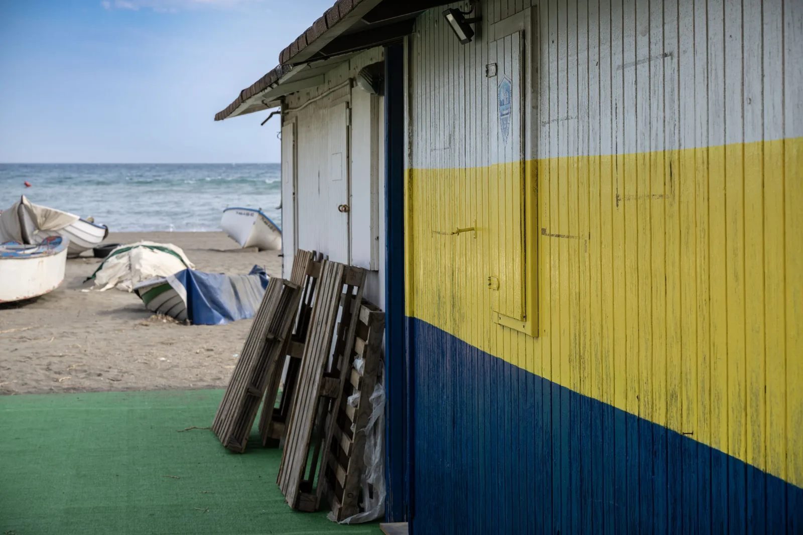 A weathered hut painted white, yellow, and blue occupies the right side, with small boats, folded wooden panels, sand, and sea beyond.