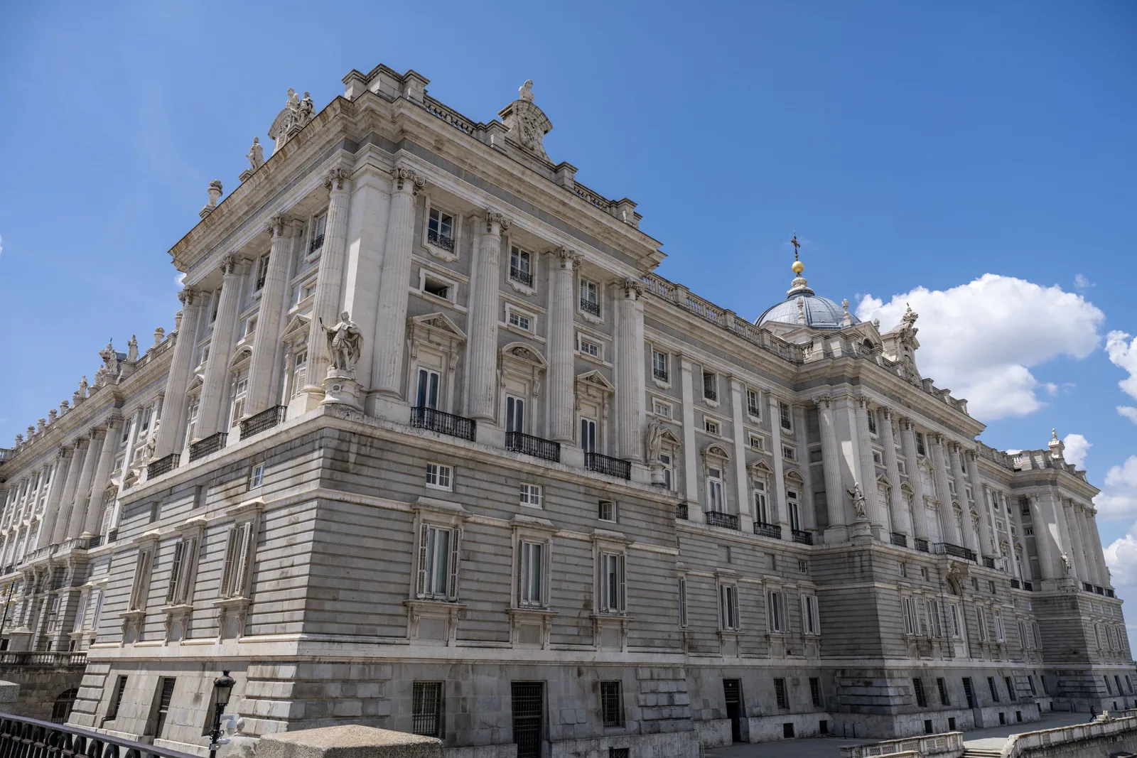 Corner view of a monumental pale stone building with tall columns, rooftop statues, multiple levels, and a dome.