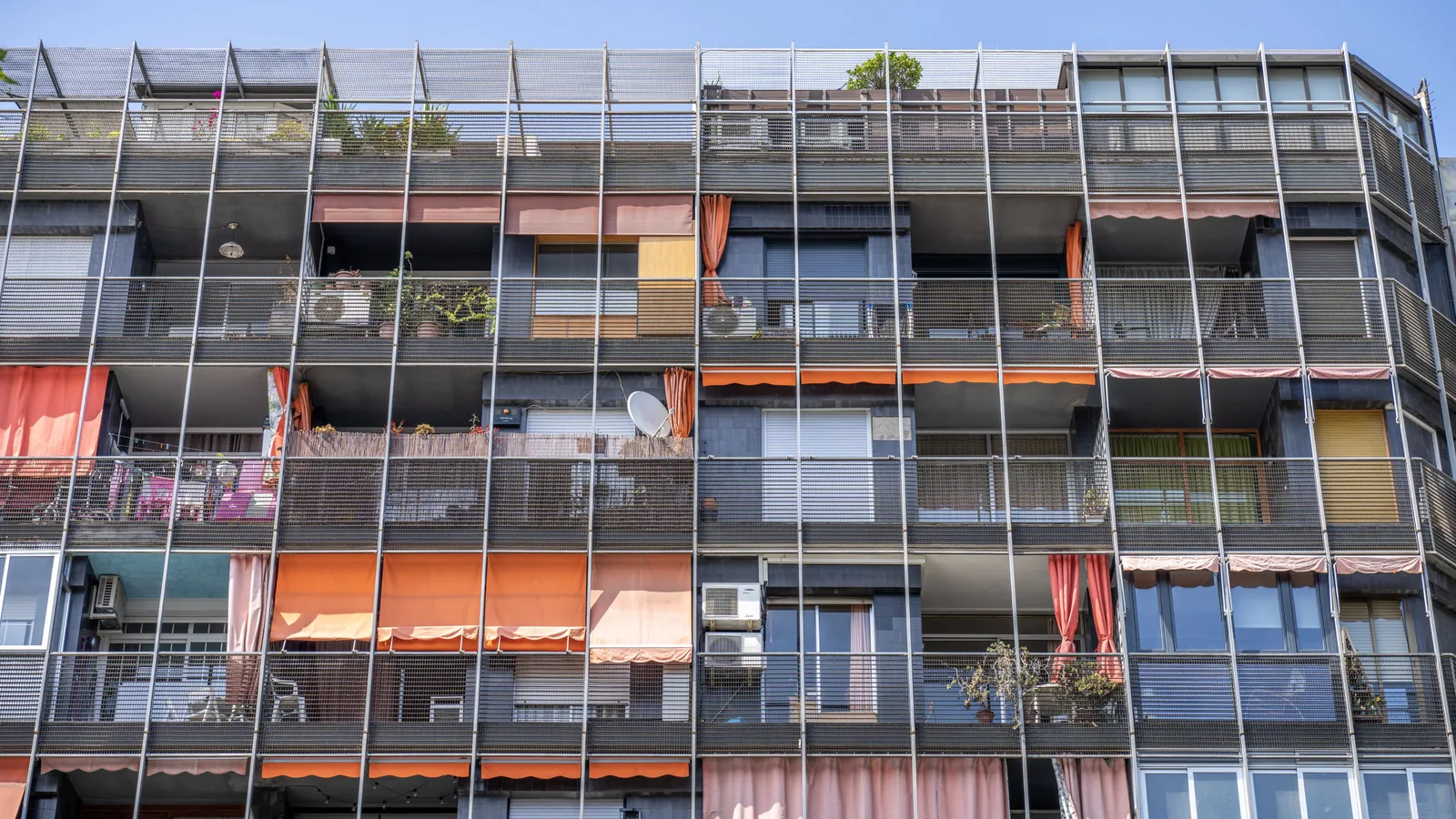 Apartment facade covered by metal balcony screens, with colorful awnings, curtains, plants, windows, and a satellite dish.