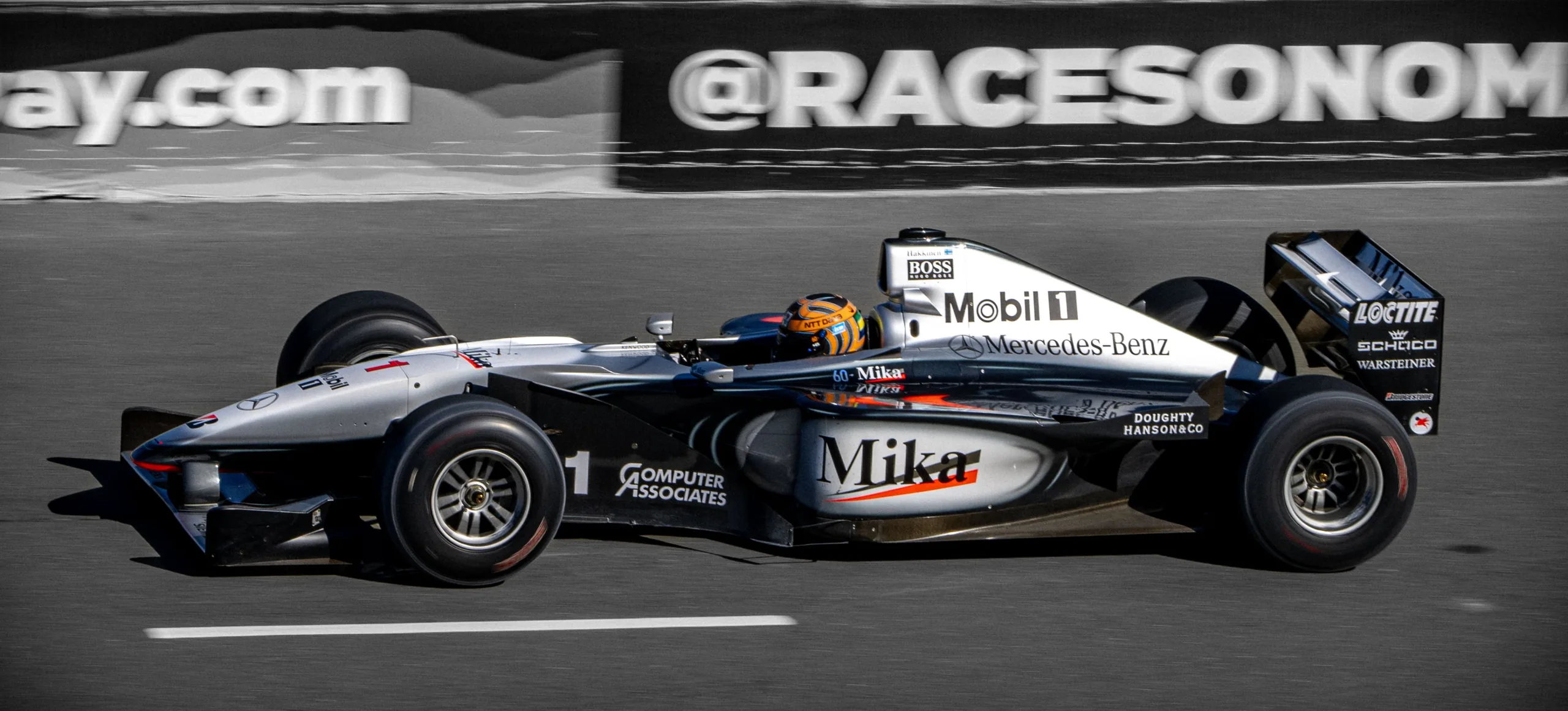 A silver-and-black open-wheel race car with Mercedes-Benz, Mobil 1, and large Mika lettering moves along a track.