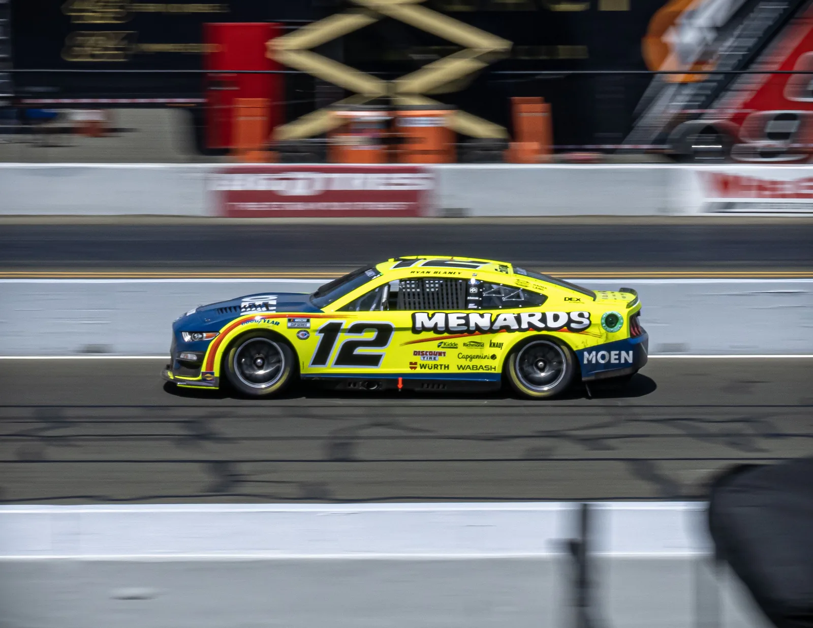 Yellow-and-blue stock car numbered 12, with Menards branding, drives along a track beside blurred pit-lane equipment.