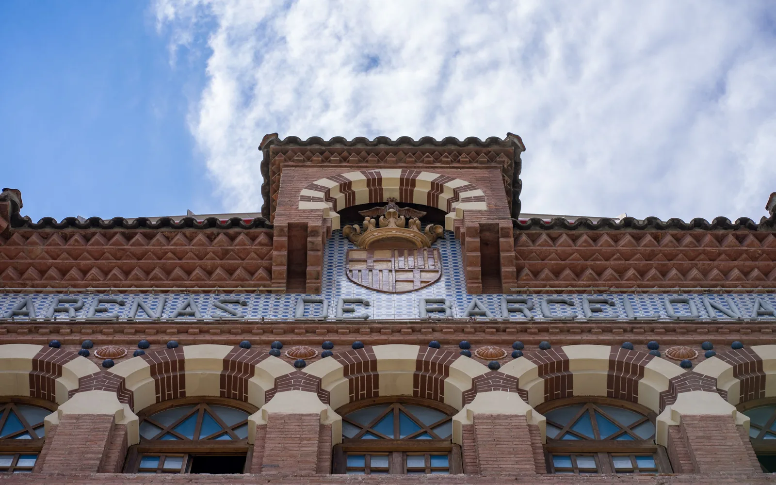 Decorated brick facade with tiled lettering, an ornamental crest, a striped arch, and repeated arches below.