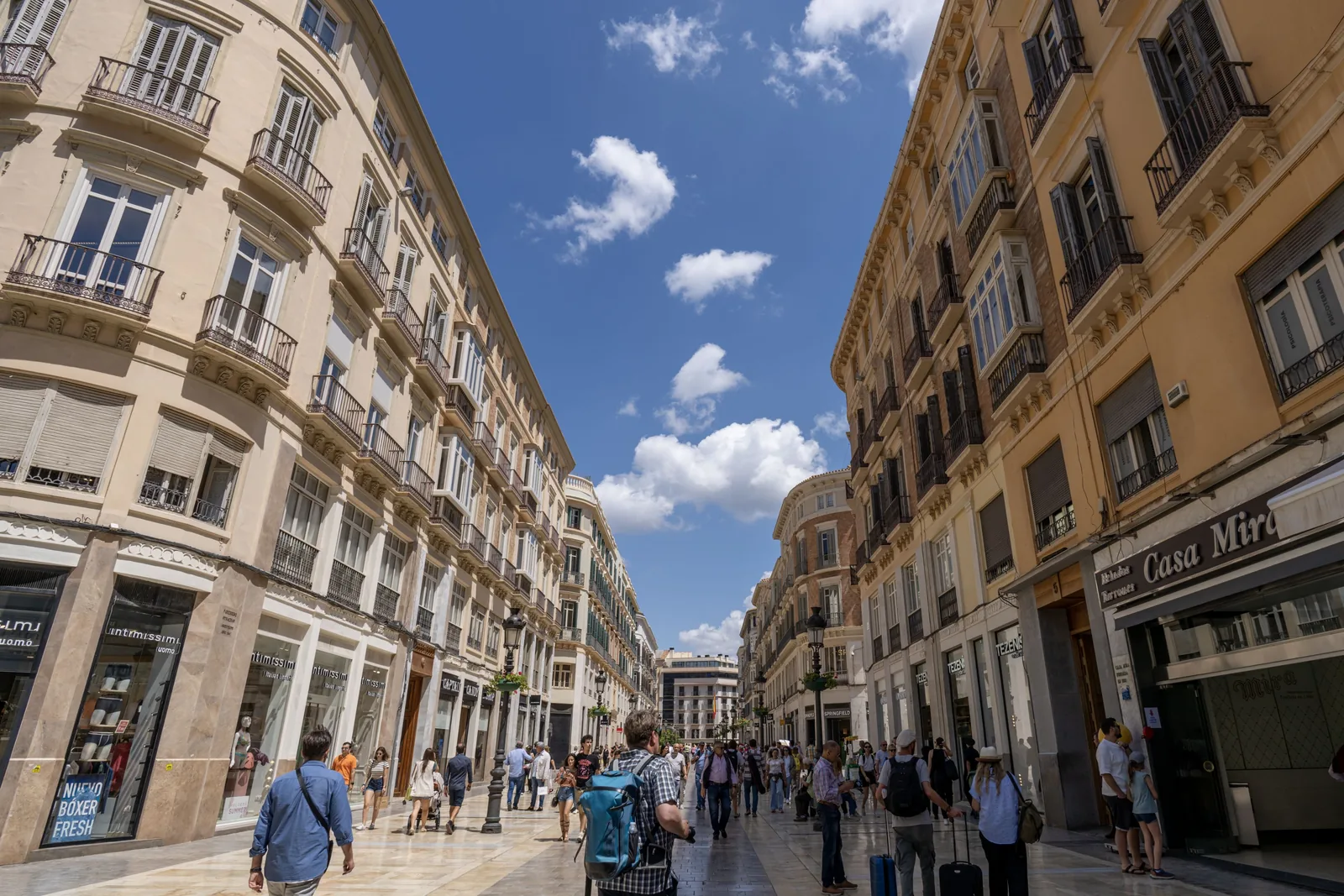 Wide pedestrian shopping street recedes between ornate buildings, with storefronts, decorative lamps, and numerous people under blue sky.