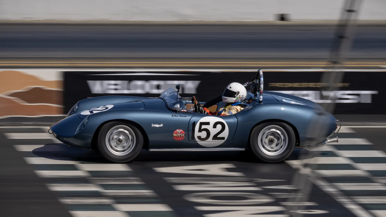 A dark blue vintage racing car marked 52 passes track barriers, with a helmeted driver visible in the open cockpit.