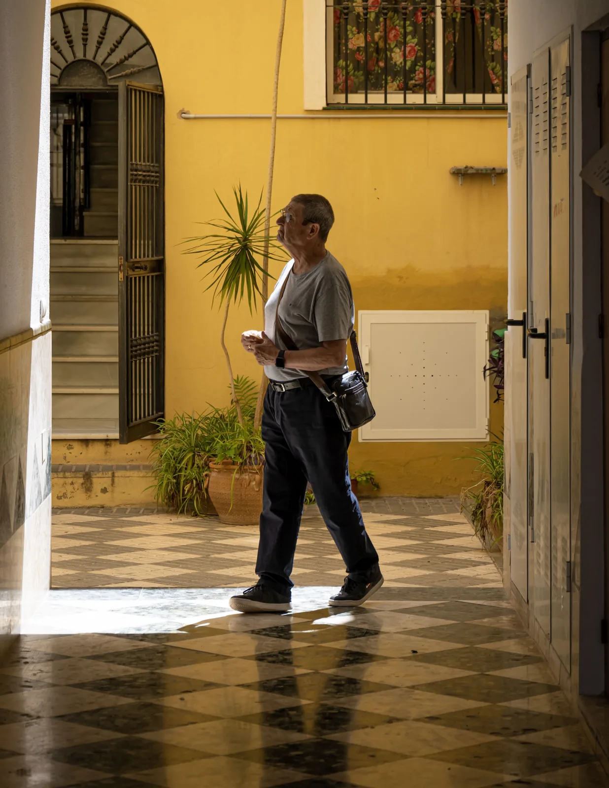 Older man with glasses and shoulder bag walks across a reflective checkered floor beside a yellow wall and potted plants.