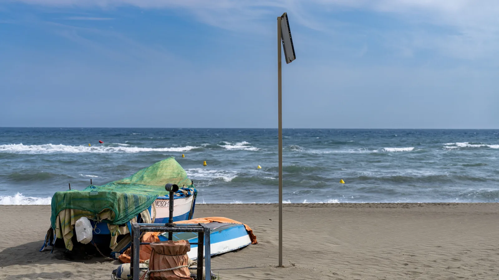 Small boats and covered beach equipment sit on sand beside choppy water, floating markers, and the back of a tall signpost.