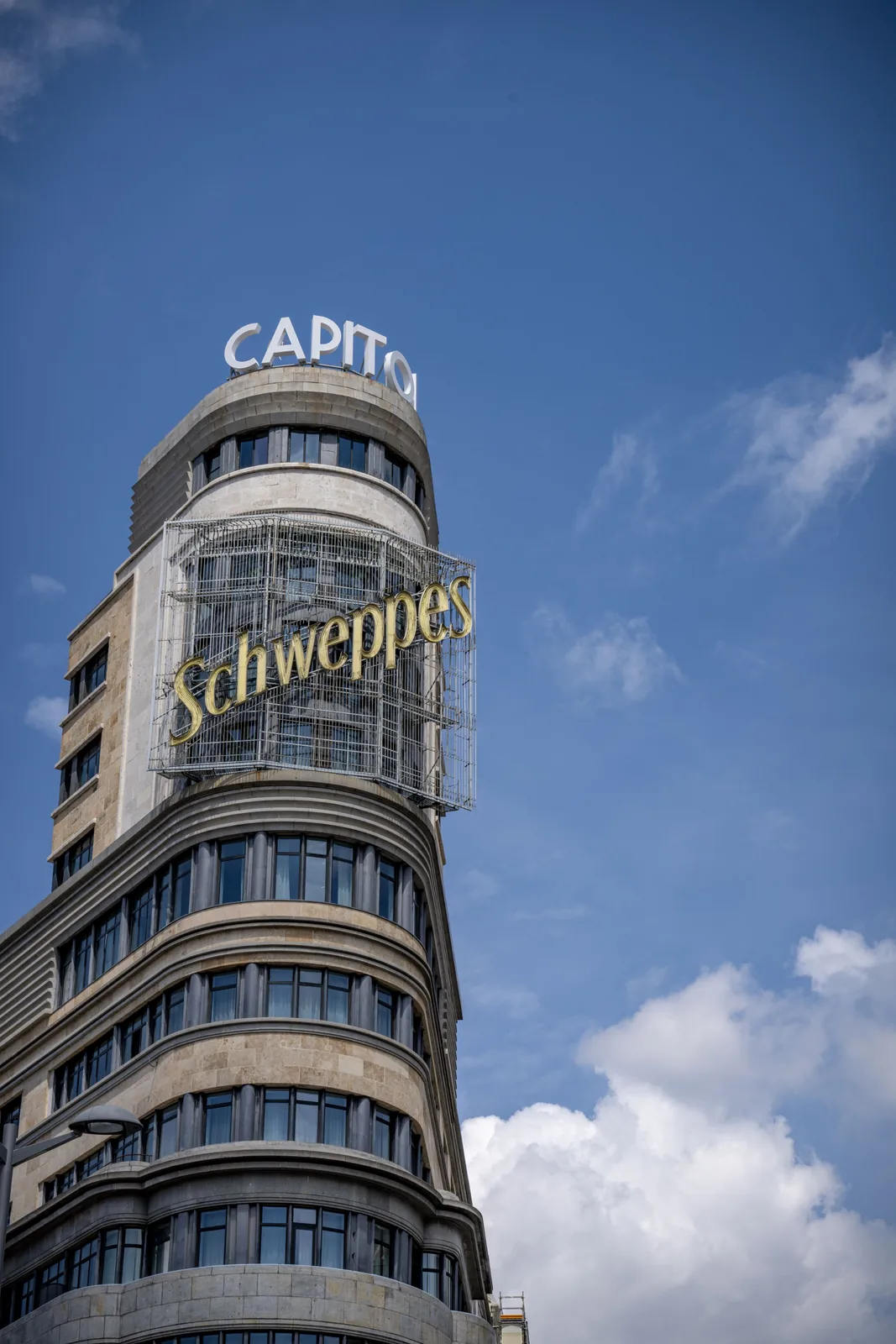 Rounded stone building rises from the lower left with large CAPITOL and Schweppes signs against blue sky and white clouds.