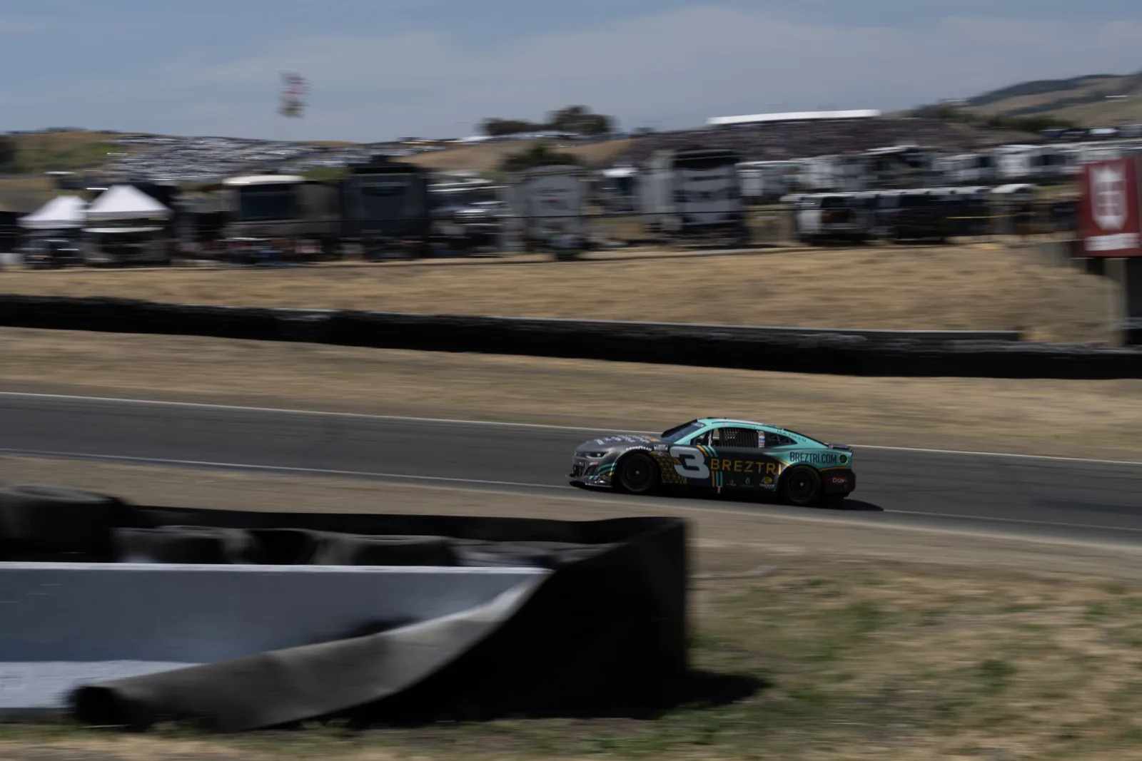 Dark stock car numbered 3 drives on a racetrack, with blurred spectators, vehicles, dry grass, and hills behind it.