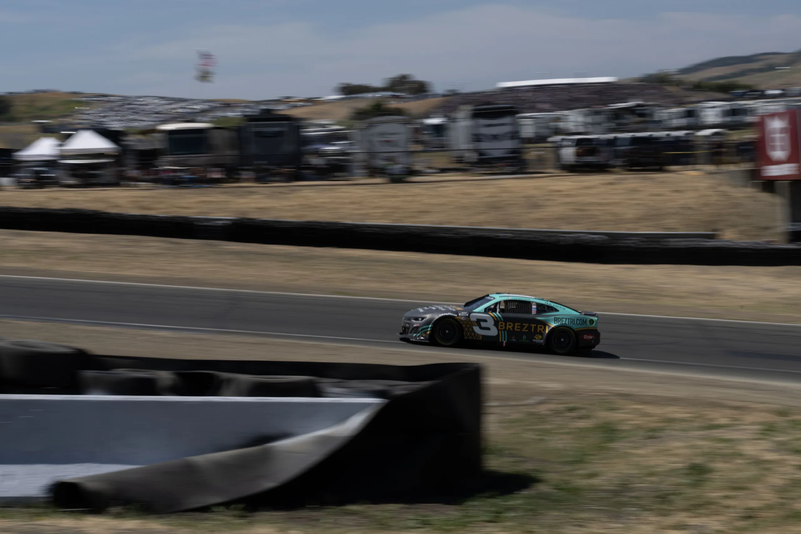 Dark stock car numbered 3 drives on a racetrack, with blurred spectators, vehicles, dry grass, and hills behind it.
