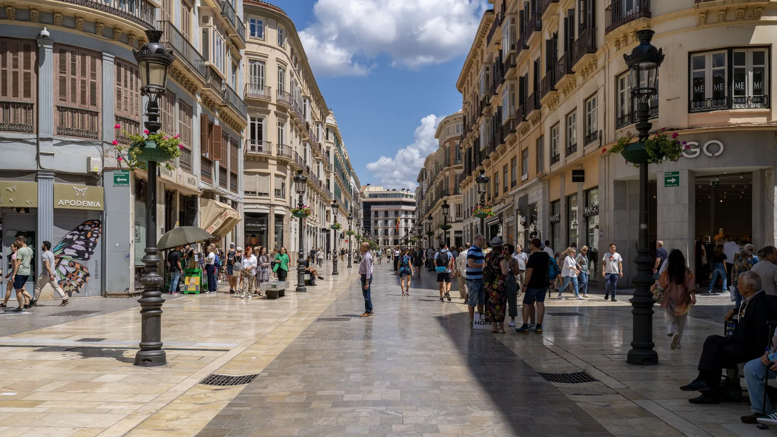 Crowded pedestrian shopping street lined with multi-story buildings, storefronts, decorative lamps, and flower planters.