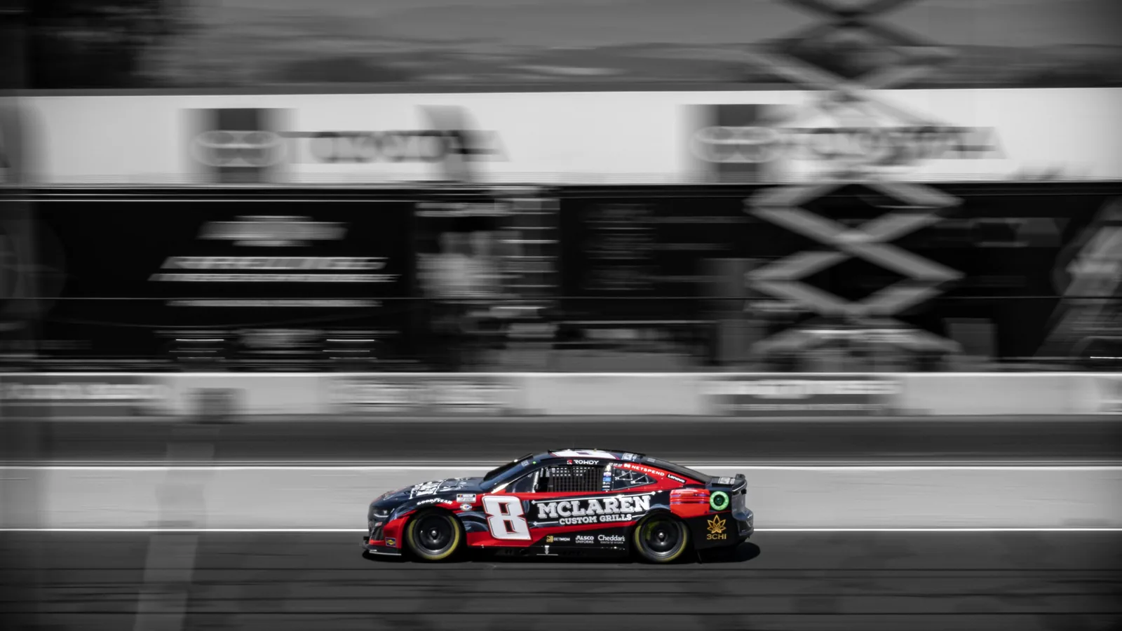 Black-and-red stock car numbered 8 drives along a racetrack, with McLaren lettering and motion-blurred pit equipment behind it.