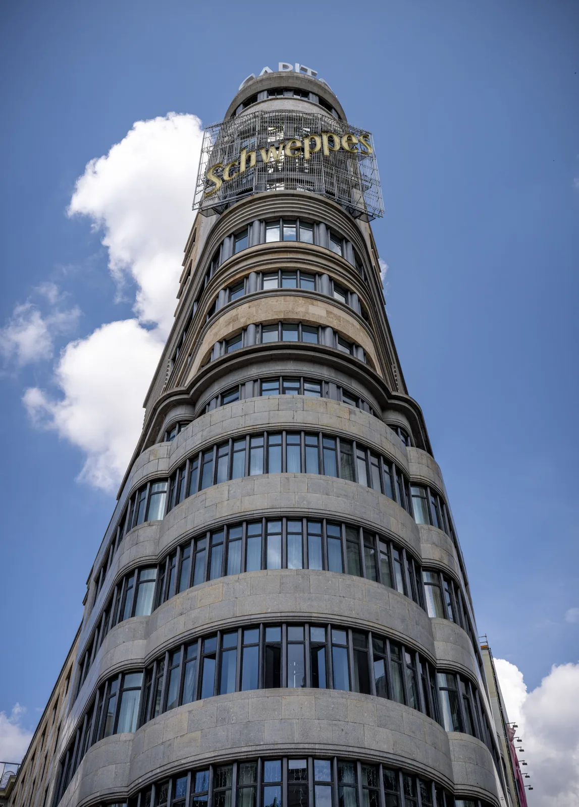 Low-angle view of a tall rounded stone building with repeated curved window bands and a large Schweppes sign.