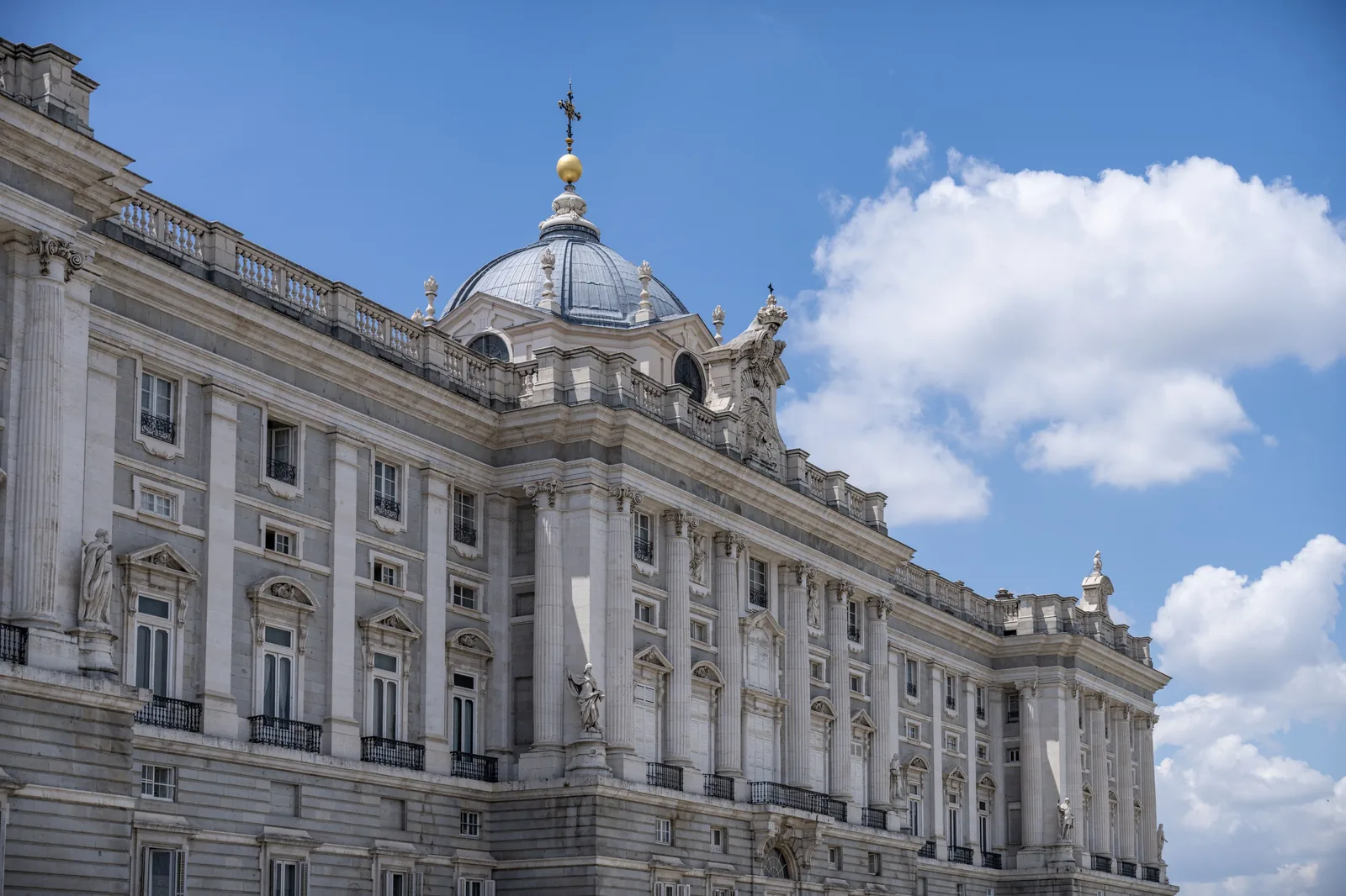 Monumental pale stone building with central dome, tall columns, statues, and long facade stands beneath blue sky and clouds.