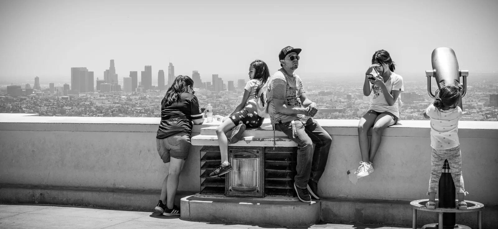 Five people gather at a rooftop overlook above a dense city skyline, including one person standing at a coin-operated viewer.