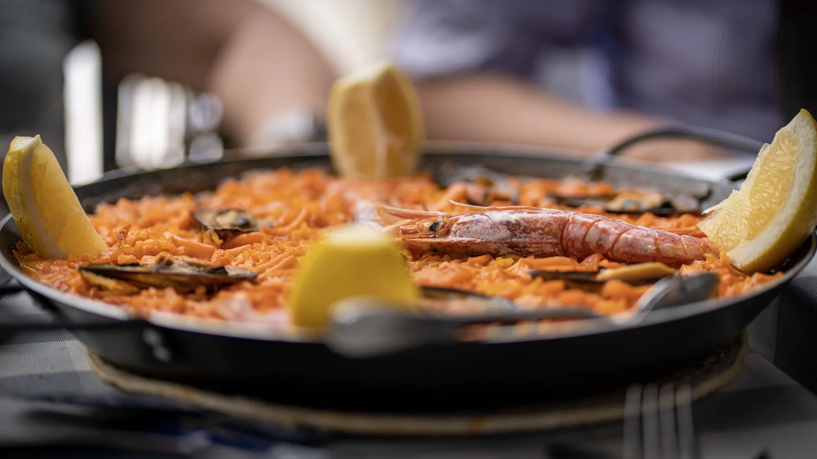 A shallow pan of orange rice is topped with a large crustacean and mussels, with lemon wedges around the rim and a blurred diner behind.