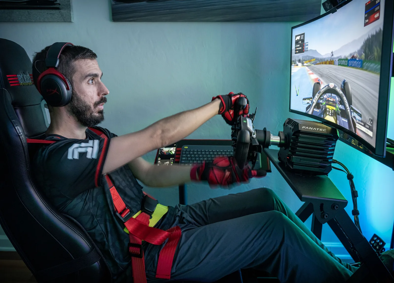 Man in racing gear operates a simulator beside a monitor showing a race car, with steering wheel, shifter, keyboard, and pedals visible.