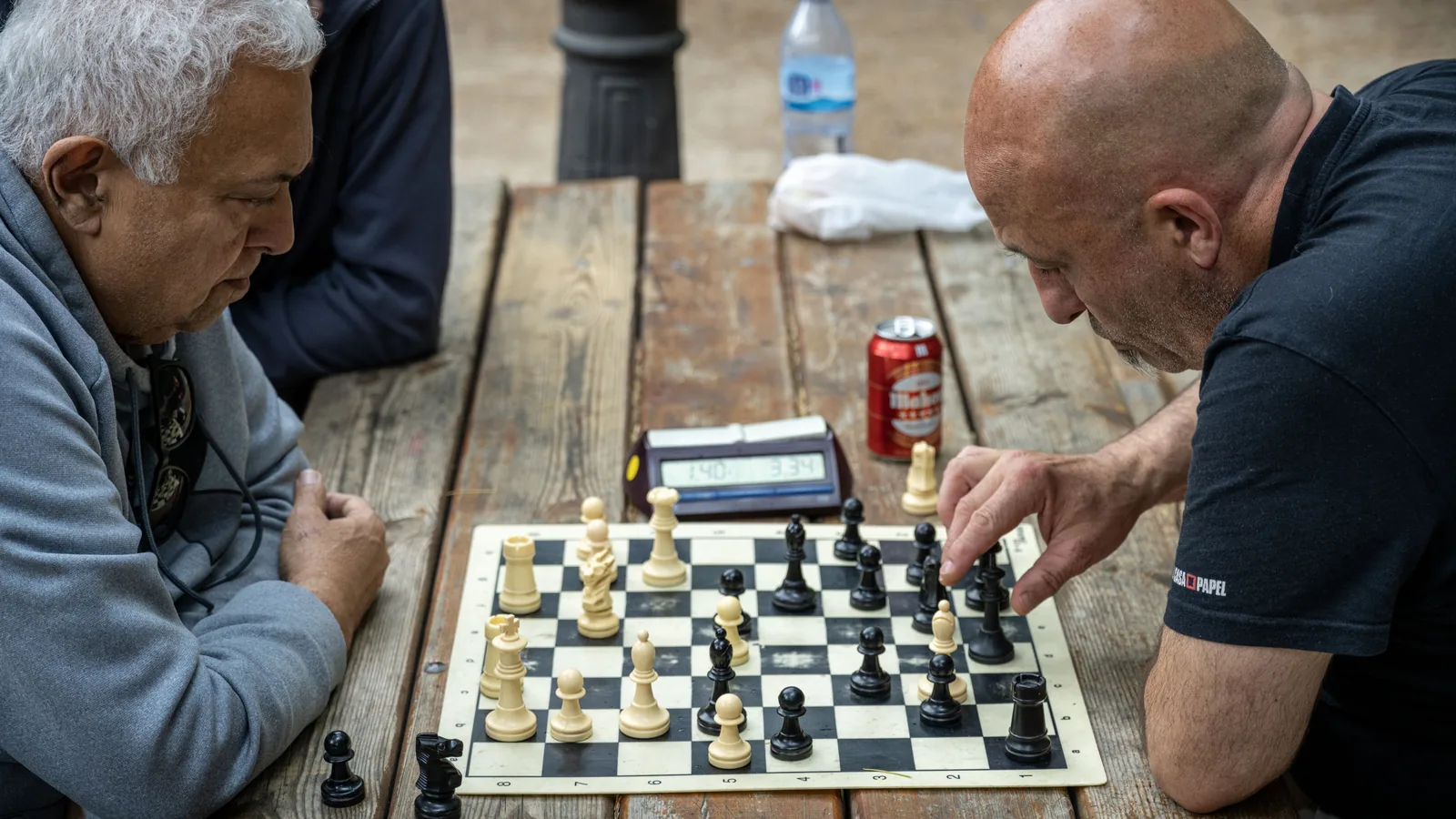 Two men study a chessboard on a wooden table, while one points at the pieces beside a digital chess clock.