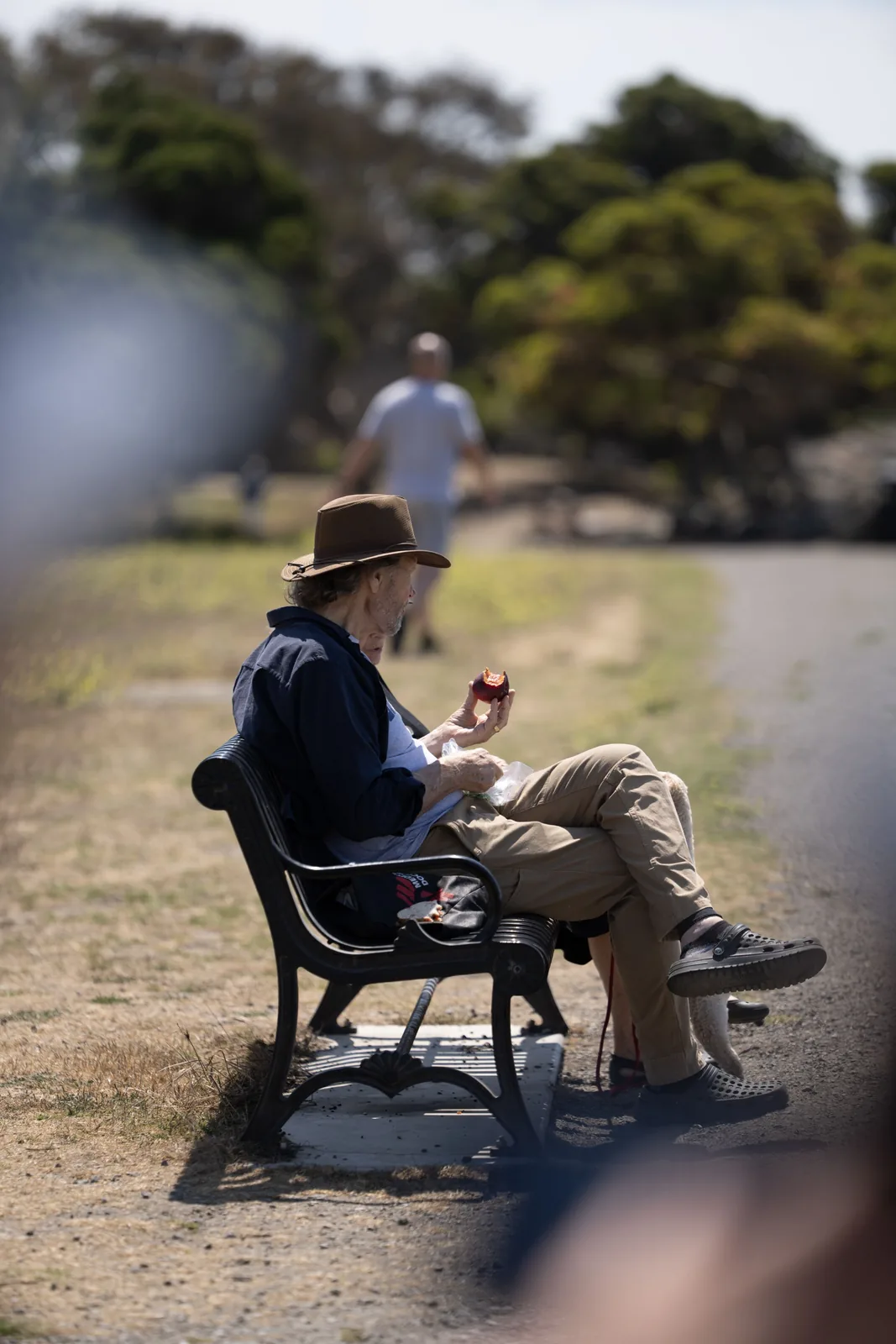 An older man in a hat sits on a park bench holding a partially eaten apple; a blurred person walks away along the path behind him.
