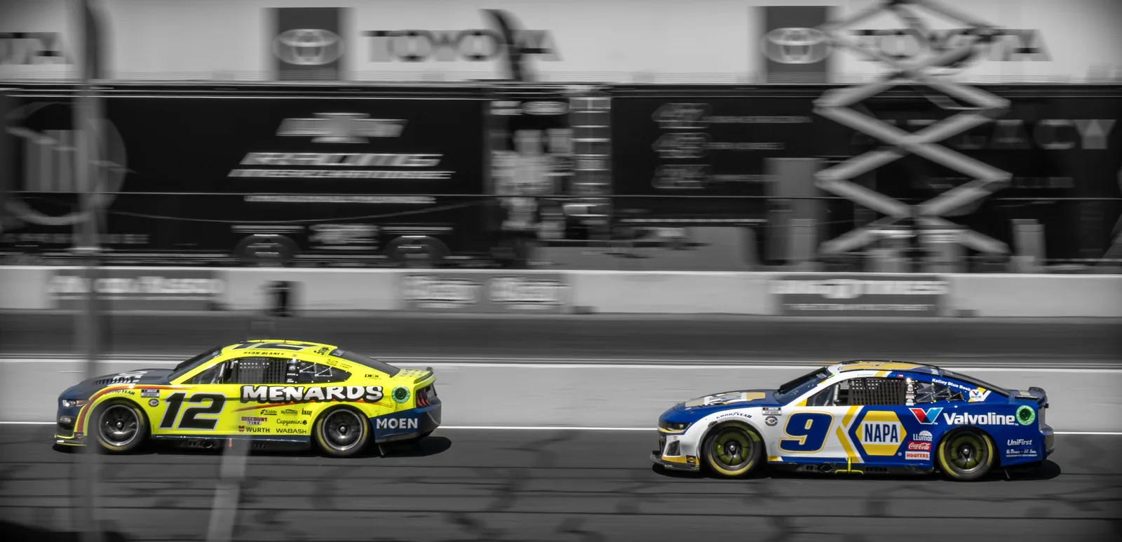Stock cars numbered 12 and 9 drive on a racetrack with blurred pit structures and signage behind them.