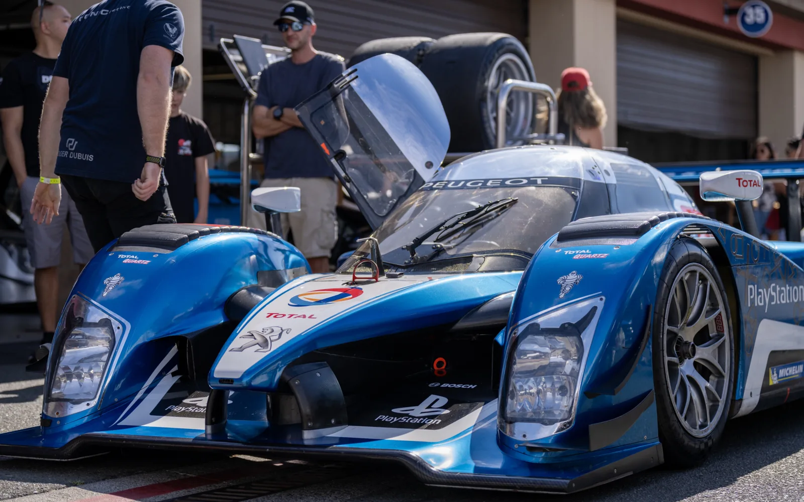 A blue-and-white Peugeot endurance racing prototype is displayed outside a garage with raised front bodywork and several people nearby.