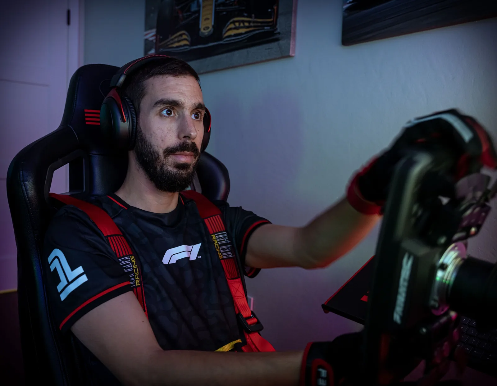 Bearded man wearing a headset and racing harness sits in a gaming chair with both hands on a simulator steering wheel.