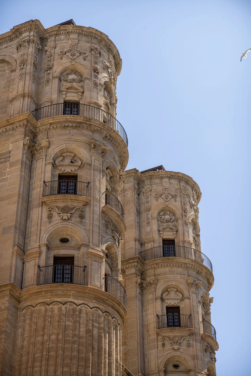 Two ornate rounded stone towers with carved panels, arched windows, and curved iron balconies rise against a clear blue sky; a bird appears at upper right.