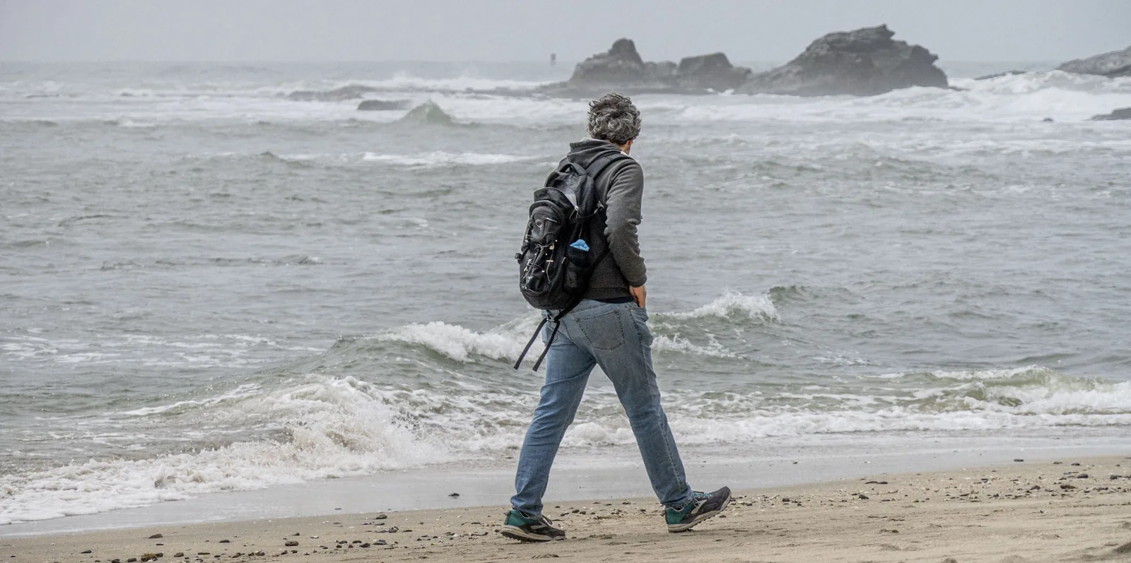 Person with a backpack walks through shallow surf beside a rocky shoreline, viewed from behind.