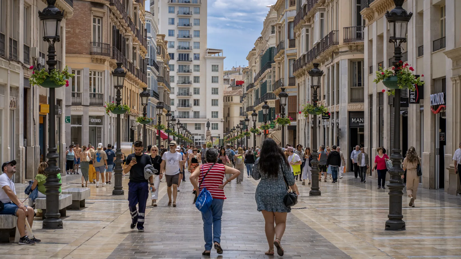 People walk along a broad paved pedestrian street lined with ornate buildings, storefronts, black lamp posts, and hanging flower planters.