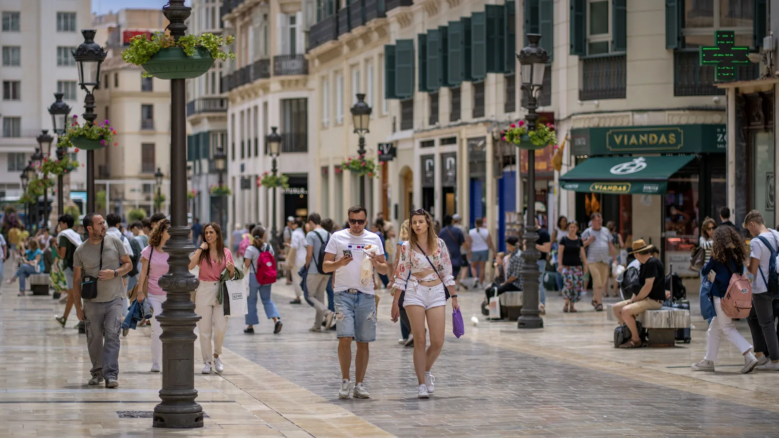Pedestrians fill a paved shopping street beside flower-filled lamp posts, ornate facades, dark shutters, and a storefront labeled Viandas.