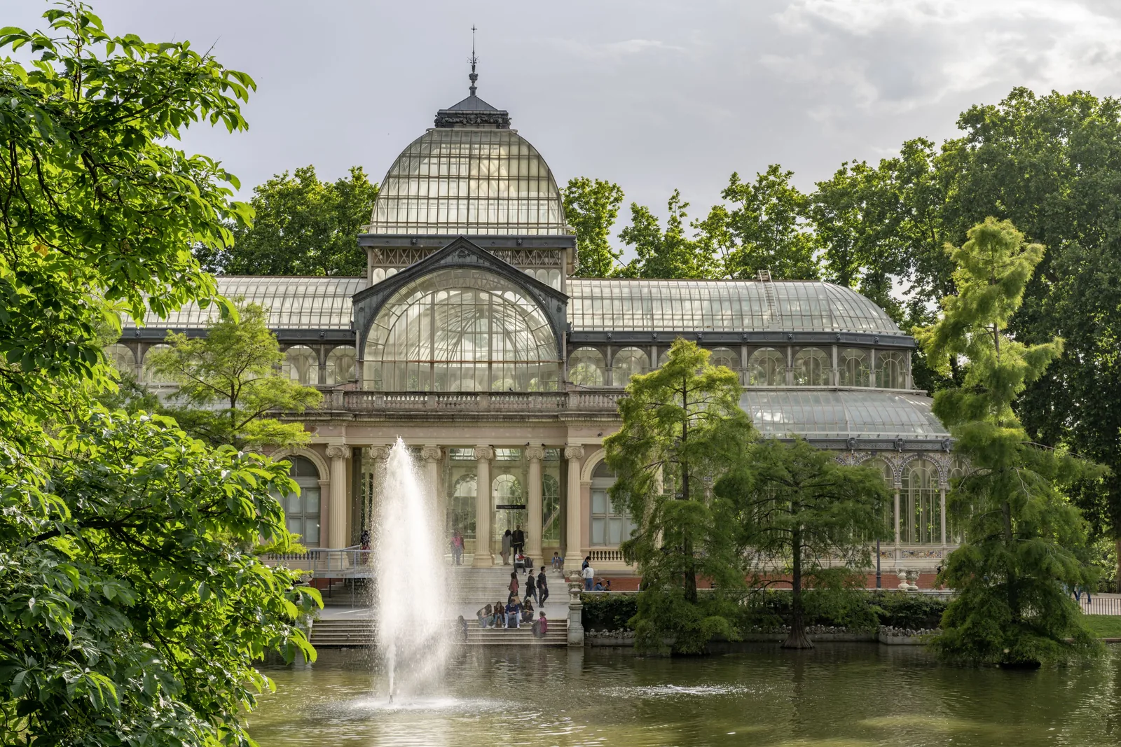 Large glass conservatory stands beyond a pond and fountain, framed by trees with people gathered on the steps.